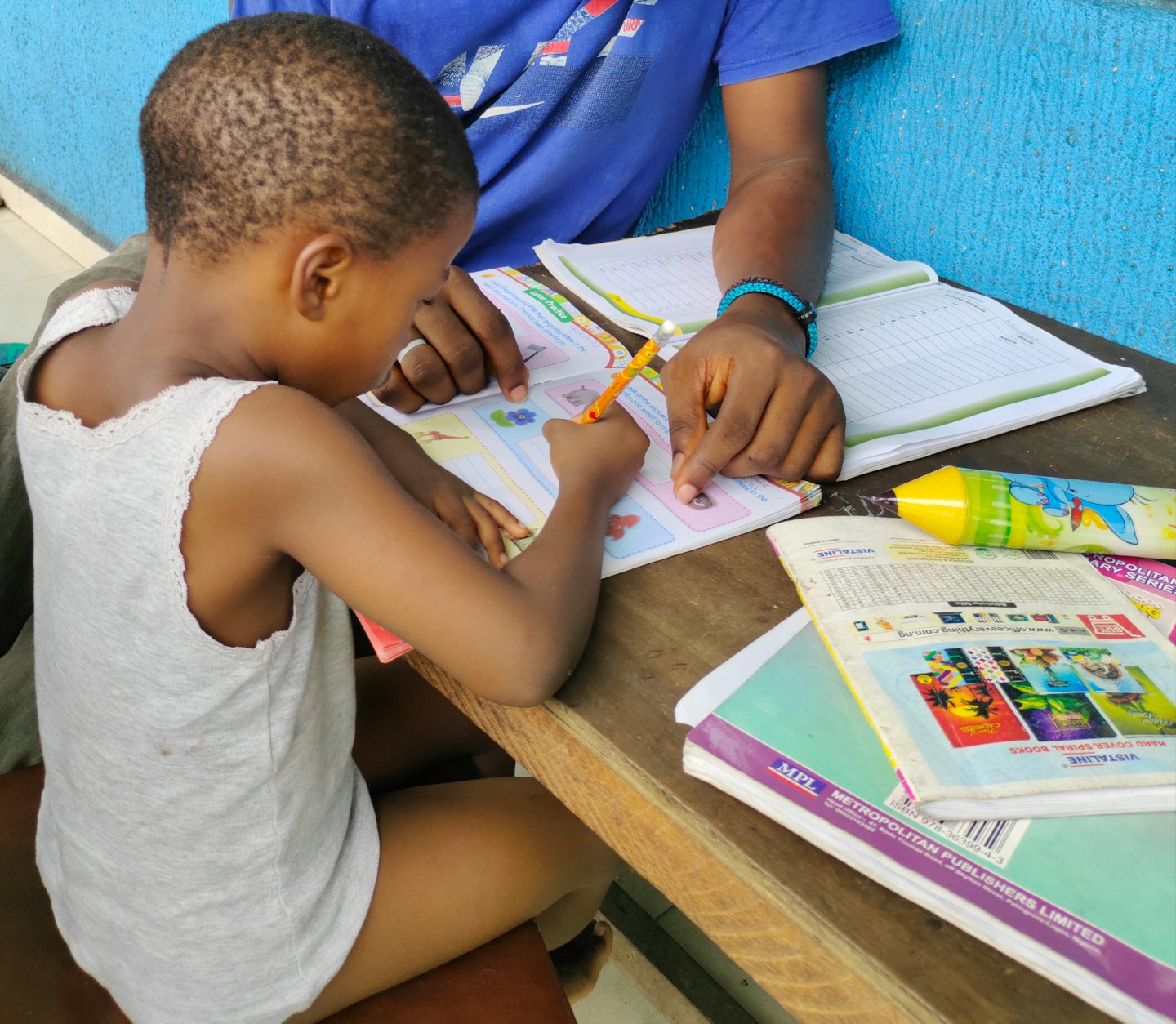 Adult guides child's hand writing in a workbook at a desk.